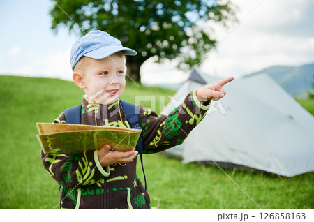 Young boy holds map and excitedly points ahead. He hiking in grassy area with tent in background, indicating camping scene. Tourist focused on navigating or planning his route. Young boy holds map and excitedly points ahead. He hiking in grassy area with tent in background, indicating camping scene. Tourist focused on navigating or planning his route. 126858163