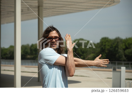 Fit handsome caucasian brunette man wearing sports casual clothes doing warm up exercise for arms on city modern outdoor public park. Summer activity. Healthy lifestyle. 126859683