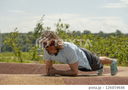 Young male person in sportswear does plank exercise to strengthen body on sport ground. Caucasian man with long curly hair enjoys training near city park. 126859686