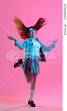 Teen girl jumping with joyful smile and flying hair in denim jacket and plaid skirt, against pink gradient background in bright neon light 126860028