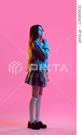 Thoughtful teen girl in plaid skirt and denim jacket standing with hand on chin against pink vibrant background in neon light 126860032