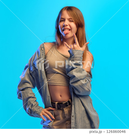 Teen girl sticks out tongue and makes rock gesture with fingers while posing in neon light against blue studio background showing bold attitude Teen girl sticks out tongue and makes rock gesture with fingers while posing in neon light against blue studio background showing bold attitude 126860046