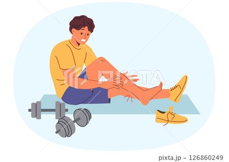 Injured sportsman sits on mat for sports near dumbbells, after stretching muscles on legs 126860249
