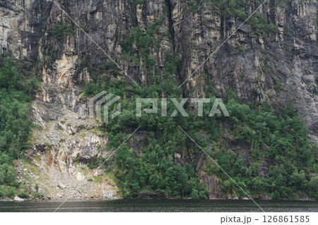 Steep rocky cliff with green forest above dark water in summer light Steep rocky cliff with green forest above dark water in summer light 126861585