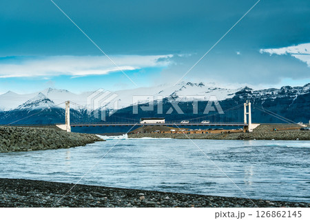 Iconic suspension bridge with car crossing over Jokulsarlon glacier lagoon and mountain range on background in Iceland Iconic suspension bridge with car crossing over Jokulsarlon glacier lagoon and mountain range on background in Iceland 126862145