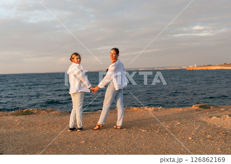 Couple Holding Hands Beach Sunset - Two people stand hand-in-hand on a beach at sunset. Couple Holding Hands Beach Sunset - Two people stand hand-in-hand on a beach at sunset. 126862169