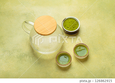 Steaming glass teapot and matcha green tea powder with two cups of prepared tea on green background top view Steaming glass teapot and matcha green tea powder with two cups of prepared tea on green background top view 126862915