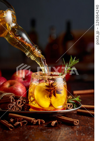 Apple cider is poured from a bottle into a glass with apple slices, anise, and a rosemary branch. Apple cider is poured from a bottle into a glass with apple slices, anise, and a rosemary branch. 126864240