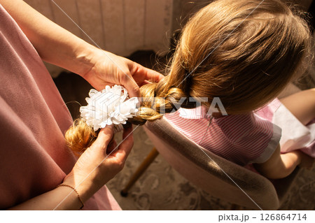 Blonde little girl sits patiently as mom carefully styles her hair into pretty braid for event. Heartwarming maternal care and childhood innocence. Blonde little girl sits patiently as mom carefully styles her hair into pretty braid for event. Heartwarming maternal care and childhood innocence. 126864714