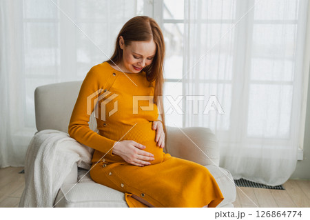 Pregnancy motherhood people expectation future. Pregnant woman with big belly sitting on chair near window at home. Girl hugging her tummy enjoying pregnancy. Maternity tenderness parenthood new life 126864774
