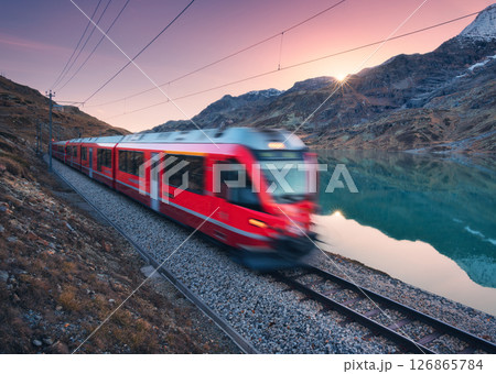 Moving red train near azure mountain lake, snowy rocks at sunset Moving red train near azure mountain lake, snowy rocks at sunset 126865784