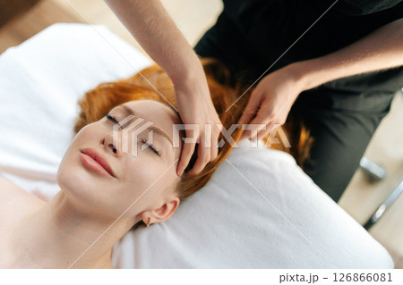 Close-up top view of skilled masseuse gently massaging temples of relaxed redhead woman with closed eyes, providing soothing head massage on comfortable massage table in spa or wellness center. 126866081
