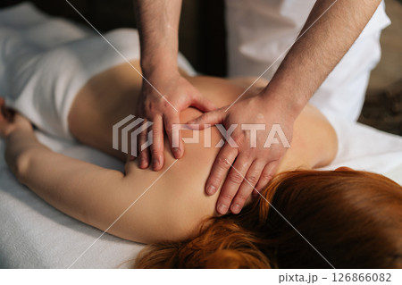 Close-up hands of unrecognizable professional masseur working on female client back, doing therapeutic massage targeting muscle tension and promoting wellness in tranquil spa environment. 126866082