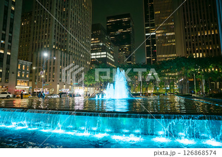 In Manhattan, a stunning fountain glows with blue light at night, surrounded by skyscrapers. People stroll along the water, enjoying the vibrant atmosphere of the city. In Manhattan, a stunning fountain glows with blue light at night, surrounded by skyscrapers. People stroll along the water, enjoying the vibrant atmosphere of the city. 126868574