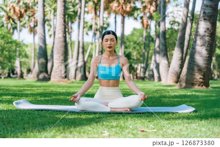 Asian woman meditating in a peaceful park setting, sitting cross-legged on green grass 126873380