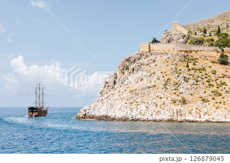 Old sailboat at the foot of a rocky coast with an ancient fortress wall on top. Beautiful seascape with a ship. Sea voyage Old sailboat at the foot of a rocky coast with an ancient fortress wall on top. Beautiful seascape with a ship. Sea voyage 126879045