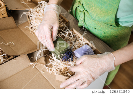 Gloved hands arrange green and lavender soaps with dried flowers in kraft packaging. Handmade business, eco-friendly packaging, natural skincare, artisan presentation 126881840