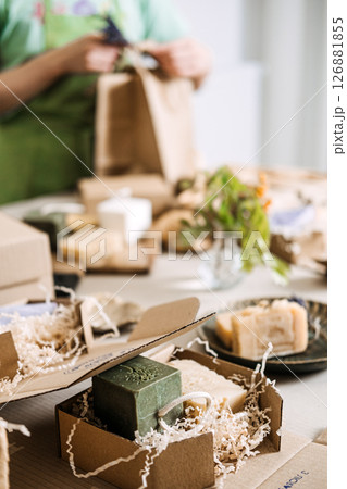 Person in green apron prepares eco-luxury soap boxes and paper bags decorated with natural elements. Zero-waste packaging, sustainable wrapping, eco-friendly branding, plastic-free materials Person in green apron prepares eco-luxury soap boxes and paper bags decorated with natural elements. Zero-waste packaging, sustainable wrapping, eco-friendly branding, plastic-free materials 126881855