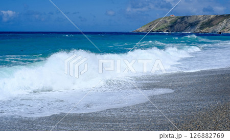 Powerful ocean waves crash onto a pebbled beach under a bright blue sky, with a rugged cliff and a distant lighthouse on a hill in the background Powerful ocean waves crash onto a pebbled beach under a bright blue sky, with a rugged cliff and a distant lighthouse on a hill in the background 126882769