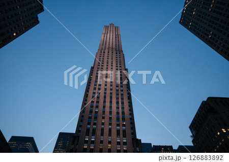 A dramatic upward view of Rockefeller Center captured at blue hour, framed by nearby skyscrapers. The fading daylight highlights the Art Deco architecture against a deep blue sky. 126883892