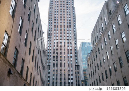 A centered view of the Rockefeller Center tower, framed tightly by symmetrical stone office buildings. The image highlights the Art Deco design and urban density of Midtown Manhattan. 126883893