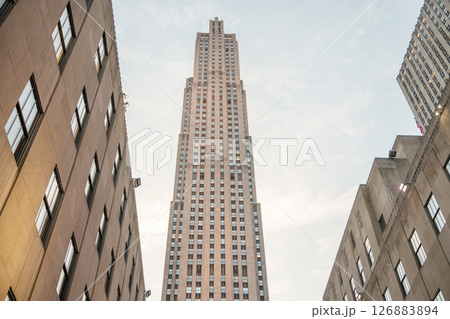 A striking view of the Rockefeller Center tower rising high between adjacent Art Deco buildings. The soft glow of the sunset illuminates the stone facade, highlighting the vertical symmetry and iconic 126883894
