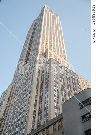 The Empire State Building towers above the street, bathed in soft evening light. Its iconic design and height dominate the skyline against a clear blue sky. 126883918