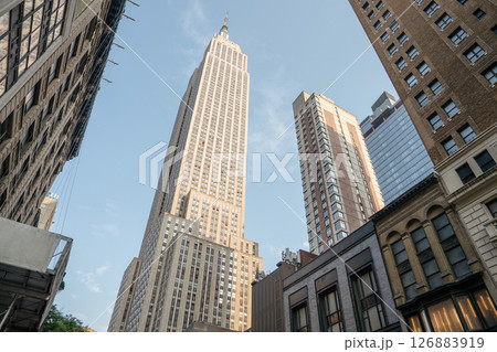 The Empire State Building towers over nearby structures in Midtown Manhattan. The clear blue sky and evening light highlight the architecture and city atmosphere. 126883919