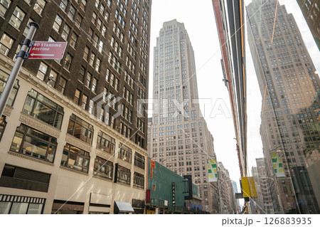 Tall buildings line a narrow street in Midtown Manhattan. One facade reflects the skyline in its mirrored glass. 126883935