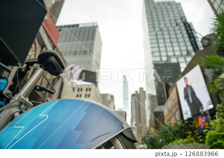 Bikes are parked on a city sidewalk in front of digital screens and tall glass buildings. The photo captures the mix of green space and high-tech transport in NYC. 126883966