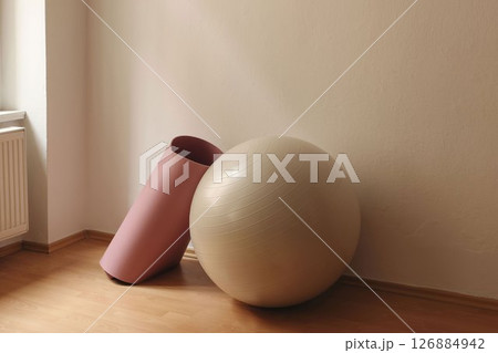 Closeup of exercise ball and pink yoga mat in soft ligh. Gym setting. Beige wall background, wooden floor. Fitness and relaxation concept. Training in studio. Set of sports equipment. 126884942