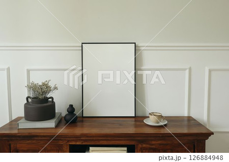 Minimalist home interior. Blank black picture frame mockup. Rustic wooden sideboard, dried flowers in a vase, and ceramic coffee cup. Neutral tones, natural light. Elegant still life. Poster display. 126884948