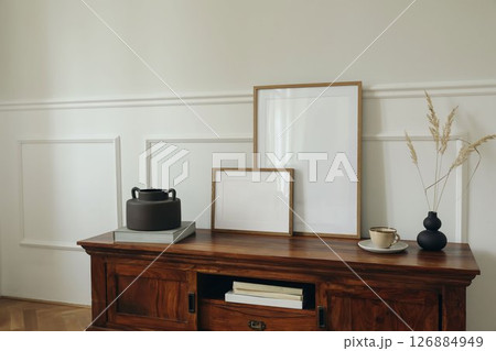 Scandinavian interior design. Modern living room. Set of blank picture mock up poster frames. Mahogany wooden dresser sideboard. Black vases with dry grass. Cup of tea, coffee. White wall background. 126884949