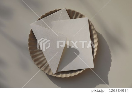 Beige fluted ceramic plate. Blank card, invitation mockup. Envelope in natural paper, softly lit by sunlight, shadows. Branding, birthday, wedding stationery. Elegant flat lay, neutral background. Beige fluted ceramic plate. Blank card, invitation mockup. Envelope in natural paper, softly lit by sunlight, shadows. Branding, birthday, wedding stationery. Elegant flat lay, neutral background. 126884954
