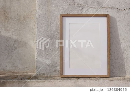 Blank thin wooden picture frame leaning against white textured wall in sunlight. Mininal elegant poster mockup. Artistic template. Natural light, soft shadows. Minimalist summer outdoor scene. A3, A2 126884956
