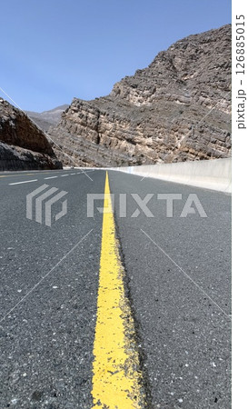 Mountain road curves sharply toward rocky tunnel, mountain road lined with cracked stripe invites passage through rugged layers, contrast between built path and raw cliffs evokes timeless tension. 126885015
