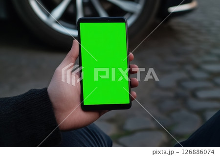 A man holds a smartphone with a green screen against the background of blurred car wheels. View from behind, hands in focus. Ideal for mockup design and advertising purposes. Copy space. Banner 126886074