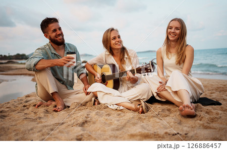 A group of friends joyfully enjoying music together while relaxing on a beach during sunset A group of friends joyfully enjoying music together while relaxing on a beach during sunset 126886457