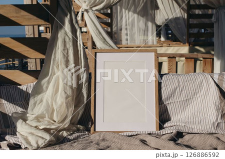 Blank wooden picture frame frame mockup. Cozy outdoor bed with striped and textured linens. Soft white curtains, natural wooden elements in warm sunlight. Summer beach vacation cabana vibes. 126886592