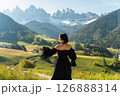 A stylish young woman in a black dress walks through sunlit fields at sunrise, with the Dolomites rising behind her. A dreamy escape into nature and elegance 126888314