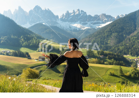 A stylish young woman in a black dress walks through sunlit fields at sunrise, with the Dolomites rising behind her. A dreamy escape into nature and elegance 126888314