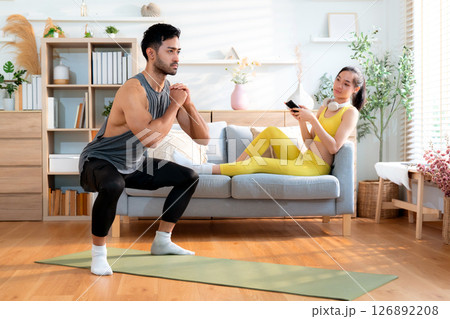 Young asian man doing squat exercise on yoga mat in living room at home while woman using phone. 126892208