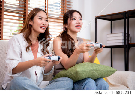 Two asian woman playing video games together with controllers in a cozy living room. 126892231