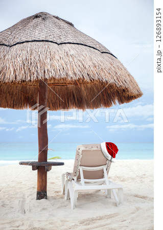 Beach umbrella and  white chair with red christmas hat on it. Santa Claus hat is a festive decoration. New Year celebration 126893154