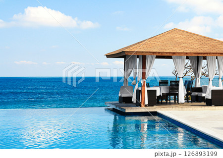 Beach with a infinite pool and a pavilion. The pavilion is covered with white curtains. The water is calm and the sky is clear 126893199