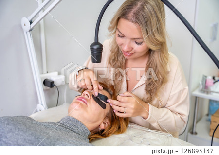 Beautician applying black eye patches before eyelash extension procedure with ultraviolet lamp. Innovative lash extension method in modern beauty salon Beautician applying black eye patches before eyelash extension procedure with ultraviolet lamp. Innovative lash extension method in modern beauty salon 126895137