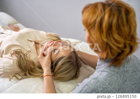 Female practitioner performs facial lifting technique on a young woman lying on a couch. Concept of energy healing, face therapy and alternative wellness practice from Access Bars. Female practitioner performs facial lifting technique on a young woman lying on a couch. Concept of energy healing, face therapy and alternative wellness practice from Access Bars. 126895150