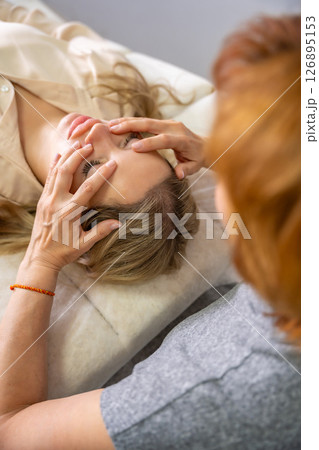 Female practitioner performs facial lifting technique on a young woman lying on a couch. Concept of energy healing, face therapy and alternative wellness practice from Access Bars. Female practitioner performs facial lifting technique on a young woman lying on a couch. Concept of energy healing, face therapy and alternative wellness practice from Access Bars. 126895153