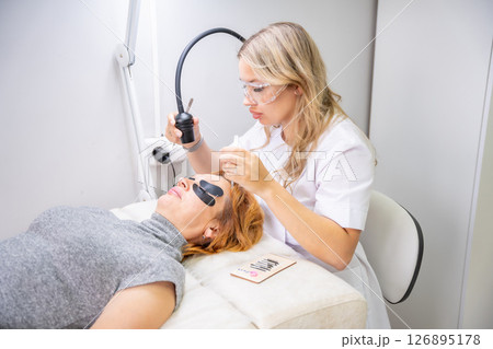 Eyelash extension procedure using ultraviolet lamp in modern beauty salon. Innovative lash enhancement technology with precise application technique. 126895178