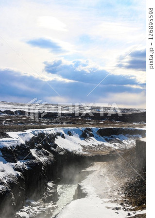 vertical photo took in Gullfoss Falls, Hvita river, Golden Circle, Reykjavik, Iceland. A waterfall cascading over rocky cliffs into a river below with snow covered areas and a clear blue sky 126895598